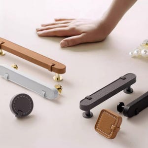 Op de afbeelding: Een assortiment kastgrepen en knoppen in verschillende kleuren, waaronder bruin, wit, grijs en zwart. De handgrepen zijn met leer omwikkeld en hebben gouden of zwarte metalen bases. Op de achtergrond is een hand te zien.