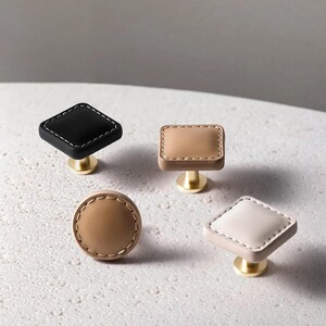 Może przedstawiać: Cztery dekoracyjne gałki do szafek o r&oacute;żnych kształtach: czarny romb, beżowy prostokąt, beżowe koło i biały romb. Każda gałka ma szytą obw&oacute;dkę i podstawę w kolorze złotym. Gałki te idealnie nadają się do dodania odrobiny elegancji meblom.