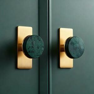 Könnte beinhalten: Zwei Schrankknöpfe mit einer goldenen, rechteckigen Rückplatte und einem runden, dunkelgrünen Marmorknopf. Die Knöpfe sind an einer dunkelgrünen Schranktür befestigt. Modernes, minimalistisches Design.