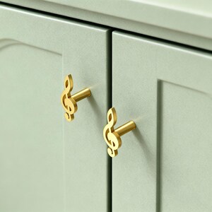May include: Close-up of two gold-colored cabinet knobs shaped like treble clefs. The knobs are mounted on a light green cabinet door. The knobs have a shiny, metallic finish, and the cabinet door has a matte finish.