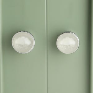 May include: Two cabinet knobs with a cloud design. The knobs have a white cloud-like pattern inside a clear, round, glass-like surface, surrounded by a silver metal rim. They are mounted on a light green cabinet door.