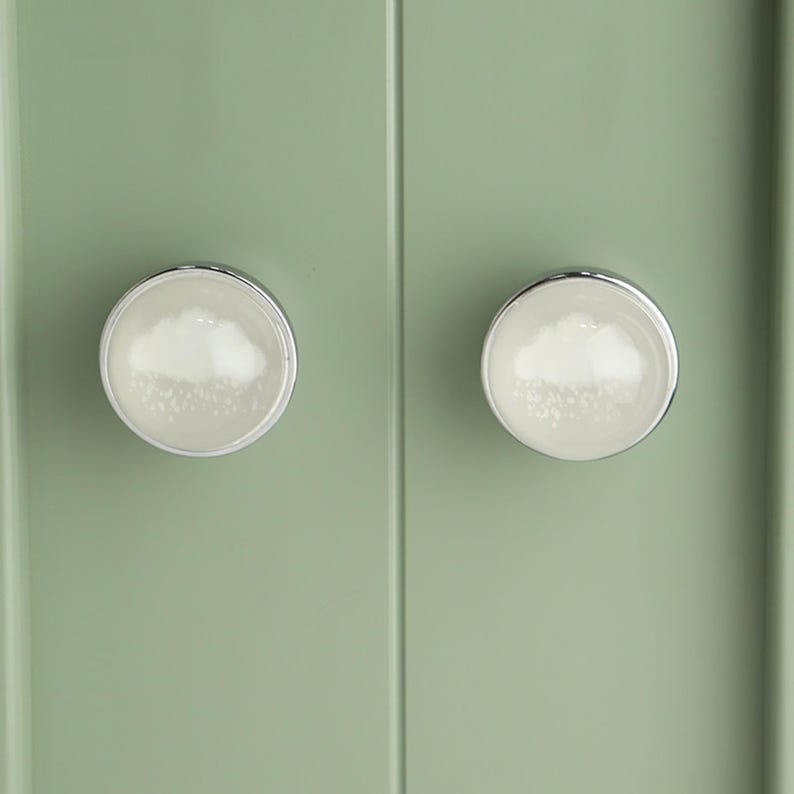 May include: Two cabinet knobs with a cloud design. The knobs have a white cloud-like pattern inside a clear, round, glass-like surface, surrounded by a silver metal rim. They are mounted on a light green cabinet door.