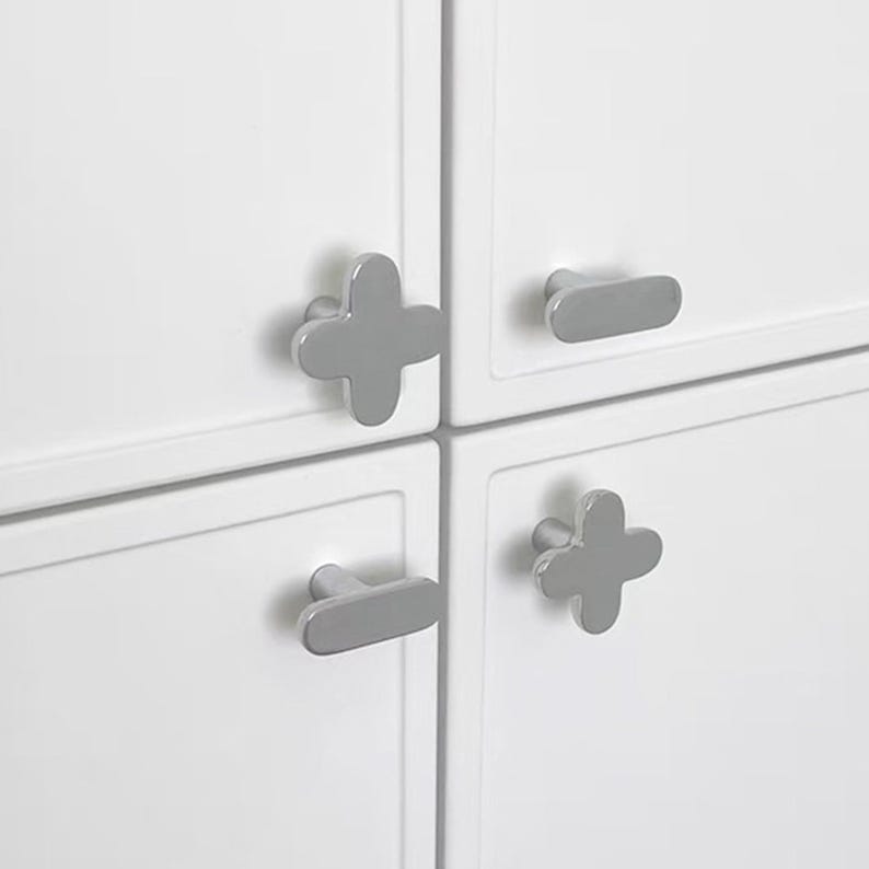 May include: Close-up of a white cabinet with four gray cabinet pulls. Two pulls are cross-shaped, and two are oval-shaped. The cabinet doors are white and have a simple, modern design.