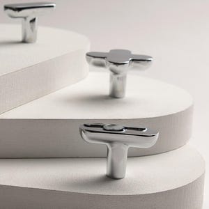 May include: Shiny, silver-toned cabinet knobs with a T-shaped design. The knobs are displayed on a white, layered surface, showcasing their reflective quality. The hardware has a modern, minimalist aesthetic, suitable for various furniture styles.