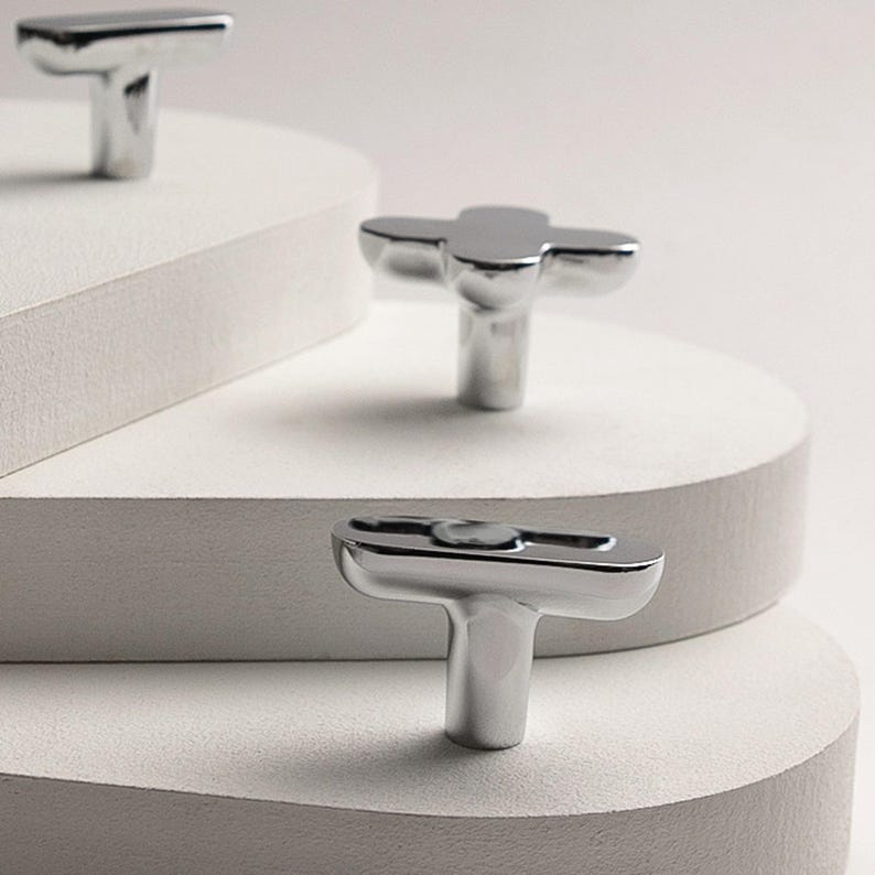 May include: Shiny, silver-toned cabinet knobs with a T-shaped design. The knobs are displayed on a white, layered surface, showcasing their reflective quality. The hardware has a modern, minimalist aesthetic, suitable for various furniture styles.