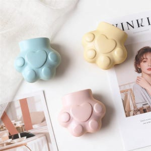 May include: Three paw-shaped ceramic containers in pastel colors: light blue, pale yellow, and pink. Each container features a heart-shaped design in the center. The containers are arranged on a white surface, with a magazine visible in the background.