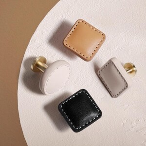 Może przedstawiać: Cztery dekoracyjne gałki do szafek w r&oacute;żnych kolorach: beżowym, jasnoszarym, białym i czarnym. Każda gałka ma kwadratową lub okrągłą powierzchnię ze sk&oacute;ry z białymi przeszyciami i złotą metalową podstawą. Idealne do mebli.