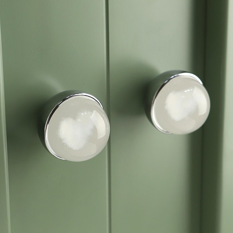May include: Close-up of two cabinet knobs. Each knob features a white, spherical center encased in a silver-colored metal base. The knobs are mounted on a light green cabinet door, creating a clean and modern aesthetic.
