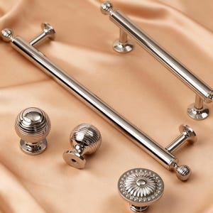 May include: A collection of polished silver cabinet hardware, including two long bar pulls, two spherical knobs with grooved detailing, and a round knob with a floral design. The hardware is displayed on a peach-colored satin fabric.