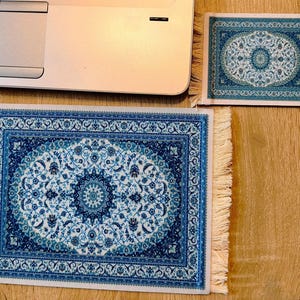 May include: Two rectangular coasters featuring a blue and white intricate floral pattern reminiscent of a traditional rug design. Each coaster has a cream-coloured fringe on the left and right sides, providing a textured finish to the edges.