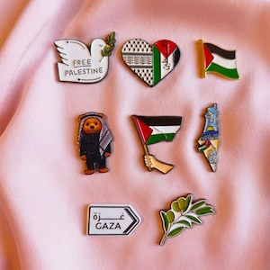 Op de afbeelding: Een verzameling van acht emaille pins, waaronder een witte duif met de tekst FREE PALESTINE, een hart met keffiyeh-patroon en Palestijnse vlag, een vlag, een beer met keffiyeh, een hand met vlag, een landkaart, een GAZA-bord en een olijftak.
