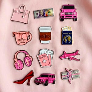 May include: A collection of twelve enamel pins featuring a pink handbag, a one hundred dollar bill, a pink SUV, a pink mug with the text coffee girlie, a coffee cup, a blue passport, pink headphones, a pink passport, a pink aeroplane, a red high heel, a pink SUV, and a stack of cash tied with a pink ribbon.