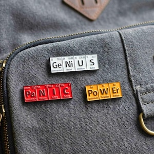 May include: Three rectangular enamel pins featuring periodic table element symbols. One white pin spells GENIUS, one red pin spells PANIC, and one orange pin spells POWER. Each pin displays atomic numbers and element names below the letters.