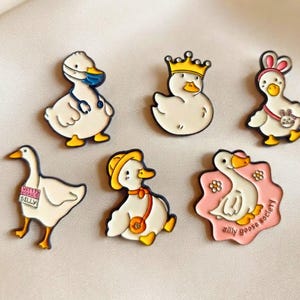 May include: Six enamel pins featuring white geese with yellow beaks and feet. Designs include a goose with a blue mask and stethoscope, a crowned goose, a goose with bunny ears, a goose with a sign, a goose with a hat, and a pink flower-shaped pin.