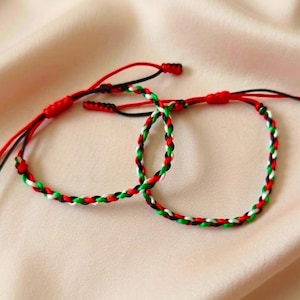 Könnte beinhalten: Zwei geflochtene Kordelarmbänder mit einem sich wiederholenden Muster aus roten, weißen, grünen und schwarzen Fäden. Jedes Armband verfügt über verstellbare Schiebeknoten aus roter Kordel, wobei lose Enden aus dem Verschlussmechanismus herausragen.