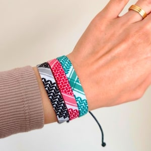 May include: Four fabric bracelets in black, white, red, pink, and teal patterns are stacked on a wrist. The bracelets have adjustable black cords. A gold ring is on a finger.