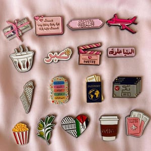 May include: A collection of enamel pins featuring various designs including a stack of cash, Arabic text, a pink aeroplane, a coffee cup, a movie clapperboard, a passport, a popcorn bucket, an olive branch, a heart with a flag pattern, and travel items.