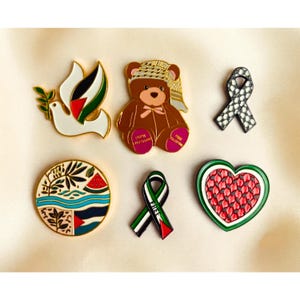 May include: Six enamel pins featuring a white dove with an olive branch, a brown teddy bear wearing a keffiyeh, a black and white patterned ribbon, a circular design with a watermelon slice, a ribbon with the colours of the Palestinian flag, and a heart-shaped watermelon slice.