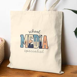 May include: A cream-coloured canvas tote bag with two shoulder straps. The front features the text "school MEDIA specialist" in a mix of cursive and block lettering, with the word "MEDIA" in blue, orange, and beige patterned fill.