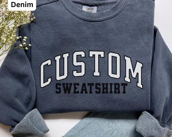 Könnte beinhalten: Ein denimfarbenes Sweatshirt mit dem Wort "CUSTOM" in großen weißen Blockbuchstaben über dem Wort "SWEATSHIRT" in Schwarz. Das Sweatshirt hat einen Rundhalsausschnitt und gerippte Ärmelbündchen. Die Farbe ist mit "Denim" gekennzeichnet.