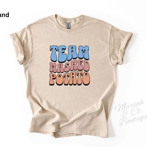 Può includere: T-shirt color sabbia con la scritta "TEAM MASHED POTATO" in un font retrò blu, rosa e arancione. La maglietta ha maniche corte e girocollo. Il nome del marchio "Mariah & Co. Boutique" è stampato in corsivo in basso a destra.