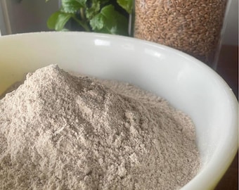 May include: A white bowl filled with light brown flour. A clear glass jar filled with brown seeds is in the background. Green leafy plants are also in the background.
