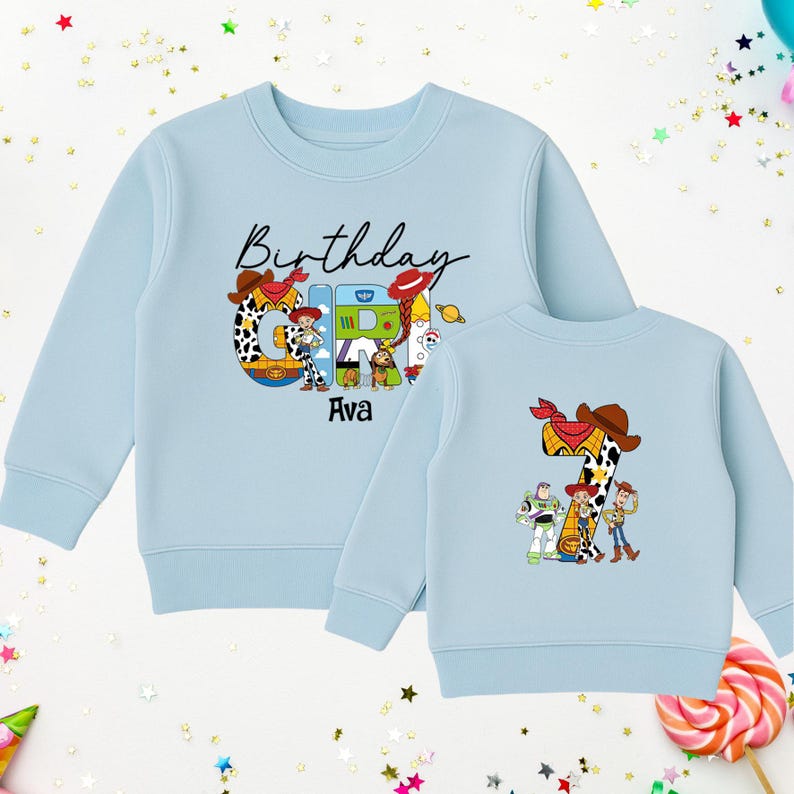 Op de afbeelding: Lichtblauw sweatshirt met een kleurrijke afbeelding met het woord "Birthday" en de naam "Ava". Het cijfer "7" wordt ook weergegeven, samen met stripfiguren. Het ontwerp wordt herhaald op de achterkant.