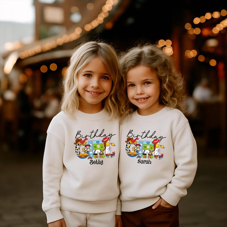 Op de afbeelding: Twee jonge meisjes dragen bijpassende cr&egrave;mekleurige sweatshirts. Elk sweatshirt heeft het woord "Birthday" in script boven een kleurrijke afbeelding met een speelgoedthema. De namen "Betty" en "Sarah" staan eronder.