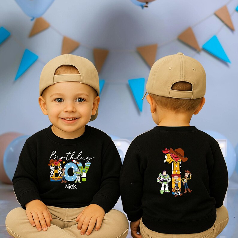 Op de afbeelding: Een zwarte longsleeve sweatshirt met een kleurrijke "Birthday BOY" afbeelding en de naam "Nick" op de voorkant. De achterkant heeft het nummer "1" en cartoonfiguren. Het kind draagt een beige baseball cap.