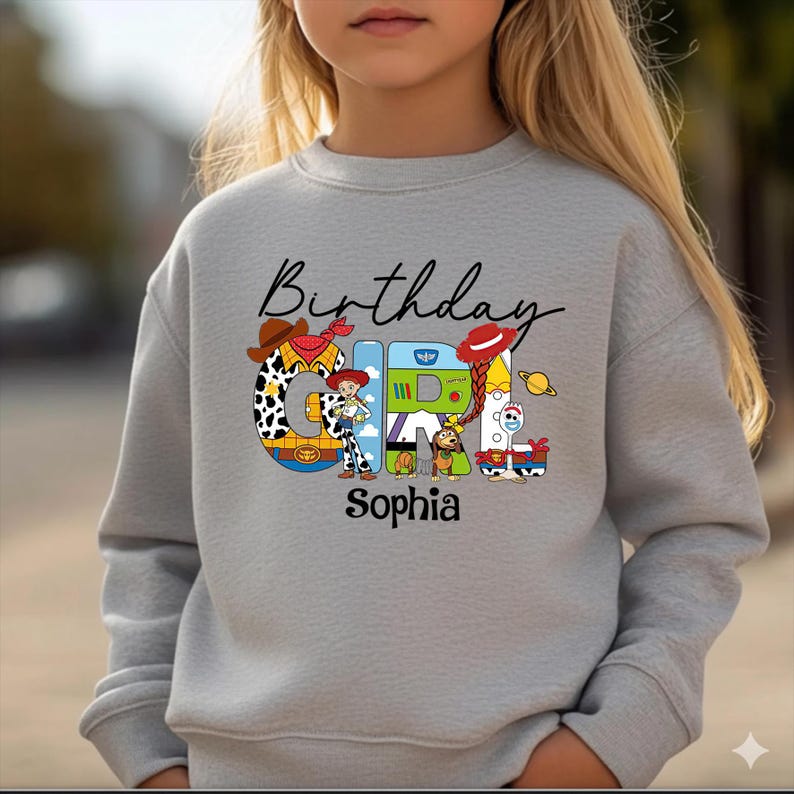 Op de afbeelding: Een lichtgrijze sweatshirt met de tekst "Birthday Girl Sophia" in zwart script. Het woord "GIRL" is ontworpen met kleurrijke cartoonfiguren en een cowboyhoed. De sweatshirt heeft een ronde hals.