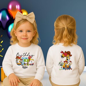 Op de afbeelding: Een witte longsleeve-trui met een kleurrijke Toy Story-afbeelding en het woord "Verjaardag" met een naam eronder. De achterkant toont een groot cijfer 2 met een cowboyhoed- en bandana-ontwerp en meer Toy Story-personages.