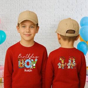 Op de afbeelding: Twee kinderen dragen rode sweatshirts en beige baseballpetten. De voorkant van het sweatshirt heeft de woorden "Birthday Boy" met een cartoonontwerp en de naam "Austin". De achterkant van het sweatshirt heeft het nummer "10" en stripfiguren.