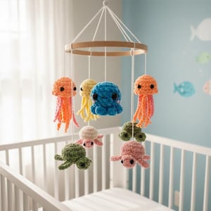 Pode incluir: Móvel de bebê feito à mão com criaturas marinhas de crochê. O móvel inclui águas-vivas laranja, um polvo azul, águas-vivas amarelas, tartarugas verdes e rosas. As criaturas penduram-se de um anel de madeira, suspenso sobre um berço branco.