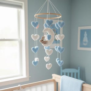 Könnte beinhalten: Handgefertigtes Babymobile mit gehäkelten blauen und weißen Herzen, einem Halbmond und einem kleinen Hasen. Das Mobile hängt an einem Holzring und ist eine bezaubernde Dekoration für das Kinderzimmer. Die Farbpalette umfasst sanfte Blau-, Weiß- und Naturholztöne.