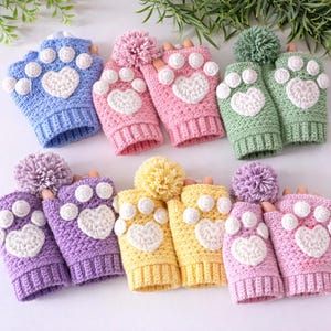 Op de afbeelding: Een verzameling kleurrijke, vingerloze gehaakte handschoenen. Elk paar heeft een pootafdrukontwerp met een hart in het midden en een pompon erop. Kleuren zijn blauw, roze, groen, paars en geel.