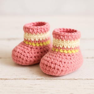 May include: A pair of handmade pink crochet baby booties. The booties have a cream and yellow striped cuff. The booties are made of a soft, textured yarn and are perfect for a newborn.
