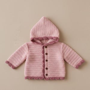 May include: A light pink crochet hooded jacket with a darker pink scalloped trim. The jacket features long sleeves and brown buttons. The hood is pointed, and the trim edges the hood and front opening.