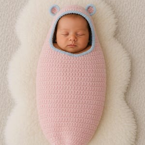 以下が含まれることがあります： 青い縁とクマの耳が付いたピンク色の手編みの繭型の寝袋に包まれた、眠っている新生児。赤ちゃんは白くふわふわのシープスキンラグの上に横たわっています。繭は赤ちゃんを暖かく安全に保つように設計されています。