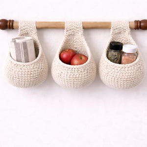 May include: Three off-white, crocheted hanging baskets suspended from a wooden rod with dark brown finials. The baskets hold a rolled towel, two red apples, and spice jars. The baskets are a decorative storage solution.