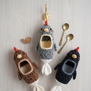 Puede incluir: Tres cestas de almacenamiento hechas a mano con forma de pollo de ganchillo en marrón, negro y gris, cada una con una cresta roja y cola blanca. También se ve un pequeño pájaro de madera y dos cucharas doradas.