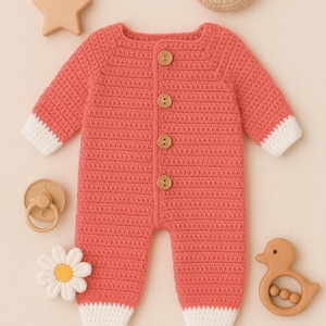 May include: A coral-colored, crocheted baby onesie with wooden buttons and white cuffs. The image also features a wooden star, a crescent moon, a pacifier, a daisy, and a duck-shaped teether, all arranged on a neutral background.