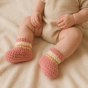 May include: A pair of handmade baby booties in a soft pink hue. The crochet booties feature a striped cuff in cream and yellow. The baby is wearing a beige onesie. The booties are designed for comfort and warmth.