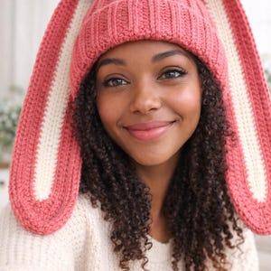 May include: A pink knitted hat with long bunny ears. The hat is a solid pink colour with cream-coloured inner ears. The hat is made of a soft, textured yarn. The hat is a fun and whimsical accessory.