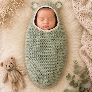 May include: A sage green crocheted baby cocoon featuring a hood with two small rounded ears and a cream-colored trim. The cocoon has a textured stitch pattern and is designed to wrap around an infant, leaving only the face visible within the hood.