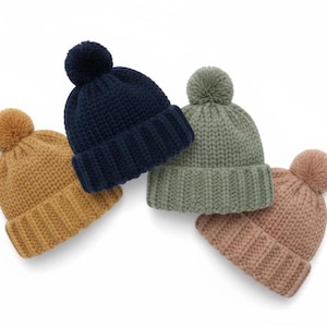May include: Four knitted beanies in mustard, navy, sage green, and dusky pink. Each hat has a ribbed brim and a matching pom-pom. The beanies are displayed on a white background.
