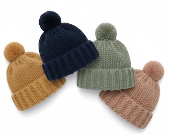 Può includere: Quattro berretti lavorati a maglia nei colori giallo senape, blu navy, verde salvia e rosa antico. Ogni cappello presenta un bordo a coste e un pompon abbinato. I berretti sono disposti su sfondo bianco.