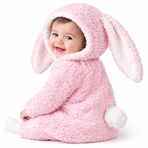 Puede incluir: Un bebé con un disfraz de conejito rosa con capucha y orejas largas, y una cola blanca y esponjosa. El disfraz está hecho de un material suave y texturizado. El bebé sonríe y lleva calcetines blancos.