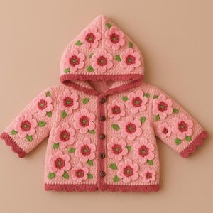 Puede incluir: Un cárdigan con capucha rosa claro para bebé adornado con grandes flores rosas tridimensionales con centros rojos y hojas verdes. El cárdigan tiene un borde festoneado y se abrocha en la parte delantera con botones marrones. La capucha también está decorada con flores.