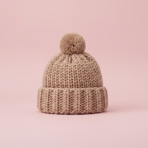 Könnte beinhalten: Beige Strickmütze mit geripptem Saum und passendem Bommel. Die Mütze ist aus dickem, strukturiertem Garn gefertigt und vor einem zartrosa Hintergrund platziert. Ein warmer und stilvoller Winter-Accessoire.