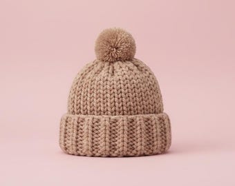 May include: A tan knit beanie with a ribbed brim and a matching pom-pom on top. The hat is made of a thick, textured yarn and sits against a soft pink background. This winter hat is a cozy accessory.
